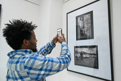 Čovjek koristi pametni telefon za fotografiranje crno-bijelih uramljenih fotografija u umjetničkoj galeriji. Posjetilac fotografira uramljene fotografije mobilnim telefonom.