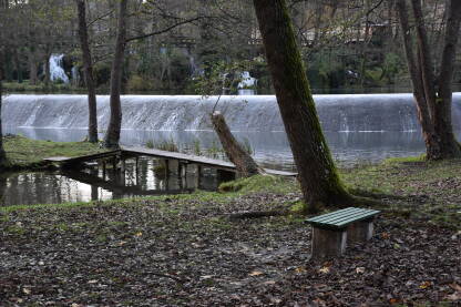 Mostić prema otoku, jezero u Jajcu Mali most sa obale na otok sa vodenom kaskadom u pozadini i zelenom klupom u donjem desnom uglu.