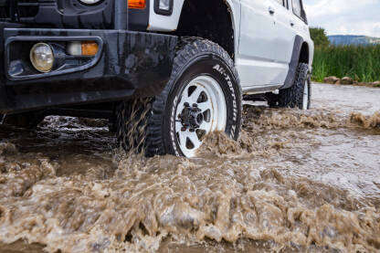 Bijelo terensko vozilo prolazi kroz plitku vodu tokom avanturističkog putovanja u ruralnom pejzažu, pod plavim nebom sa oblacima.