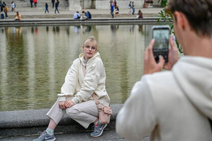 Djevojka pozira na ulici, plavuša sa naočarama se fotografiše Beč, Austrija: dečko fotografiše devojku svojim mobilnim telefonom. Turisti se fotografišu ispred crkve i fontane u gradu. Crkva sv. Charlesa.