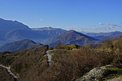 Asfaltni vijugavi put, prema selu, na koti iznad kotline. U pozadini se vide vrhovi brda i planina. ​