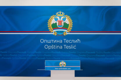 Grb Opštine Teslić na plavoj pozadini sa latiničnim i ćiriličnim natpisom Opština Teslić. U prvom planu govornica sa grbom Opštine Teslić. Press sala.