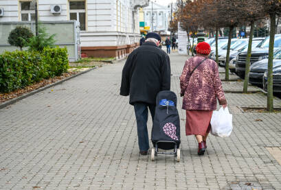 Stariji par hoda trotoarom s kolicima za kupovinu na točkićima. Stariji par odlazi ulicom. Stariji par s kolicima za kupovinu na pločniku. Penzioneri se vraćaju kući nakon kupovine.