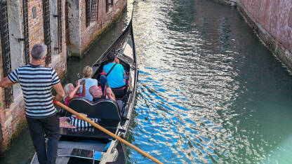 Gondola sa turistima u Veneciji Venecija, Italija: Gondola na rijeci. Istorijske građevine uz riječni kanal. Popularna turistička destinacija. Turisti istražuju grad.