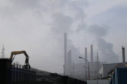 Industrijalizacija i zagađenje u Tuzli U dubini kadra su dimnjaci, kompleks termoektrane u Tuzli, vidljiv u naznakama kroz smog i maglu. Lijevo u kadru iza ograde je prometnica, u trenutku prolazi autobus i izvode se građevinski radovi.