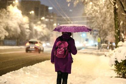 Žena sa kišobranom šeta gradom dok pada snijeg zimi. Pahuljice snijega.
