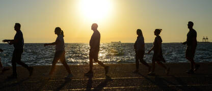 Silueta ljudi dok šetaju rivom uz more. Grupa ljudi u pokretu. Osobe šetaju uz more tokom zalaska sunca.