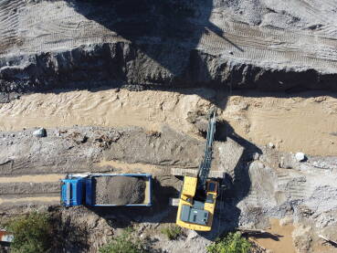Bager utovara materijal na kamion, snimak dronom. Bager Bager utovaruje materijal na kamion, snimak dronom. Bager radi u rijeci nakon velikih poplava. Žuti bager kopa u rijeci.