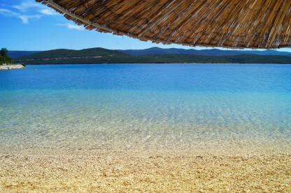 Plaža Moračna (Klek, Hrvatska) sa plavim morem, suncobranom od slame i šljunčanom plažom.