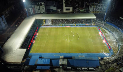 Fudbalski stadion Grbavica tokom fudbalske utakmice noću, snimak dronom. Fudbalski stadion tokom fudbalske utakmice noću, snimak dronom. Igrači na terenu. Zeleni sportski teren, svjetla i tribine na stadionu. Stadion Grbavica, Sarajevo.