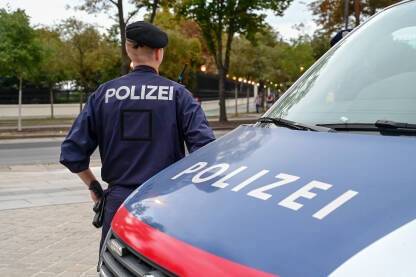 Policajac stoji uz policijski auto. Policajac u uniformi. Policajac stoji uz policijski auto u Beču, Austrija. Policajac u uniformi patrolira ulicom. Simbol policije na vozilu i uniformi.