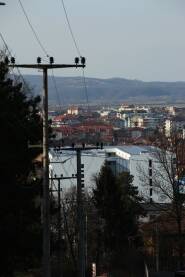 Pogled na grad sa puta Pejzaz grada i planine, stuboci za elektricne zice sa strane, drvece, zgrade, gradjevine, stanovi
