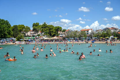 AK1_9197 Grupa ljudi se sunča na plaži i kupa u Jadranskom moru. Turisti uživaju i zabavljaju se na plaži. Prepuna plaža. Turizam.