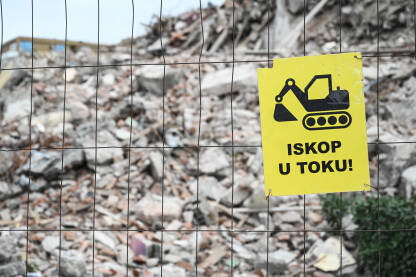 Znak na gradilištu: Iskop u toku. Znak na gradilištu: Iskopi u toku. Ograda ispred gradilišta. Srušena zgrada sa gomilom betona i cigli u pozadini.