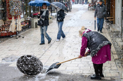 Žena koja čisti snijeg na Baščaršiji tokom padavina. U pozadini se vide prolaznici sa kišobranima, prodavnice i mokra kaldrma.