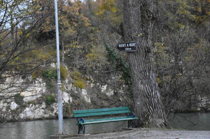Usamljena klupa i znak rent a boat (iznajmljivanje čamaca) Zelena klupa pored drveta sa jezerom u pozadini u kasnu jesen.