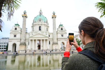 Djevojka fotografiše crkvu u Beču Beč, Austrija: Djevojka fotografiše crkvu svojim mobilnim telefonom. Turisti fotografišu ispred crkve i fontane u gradu. Crkva sv. Charlesa.