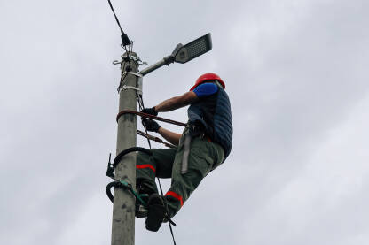 Električar na banderi Električar radi na rasvjetnom stubu. Osoba nosi zaštitnu kacigu, prsluk i rukavice. Pozadina je oblačna.