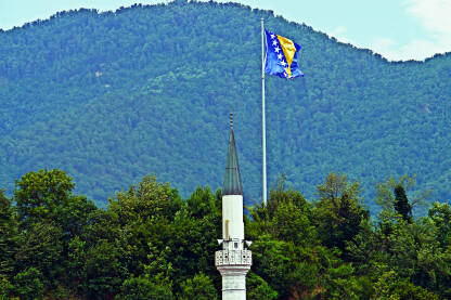 Munara stare džamije, u Podbrežju, na ulazu u Jablanicu. U pozadini na brijegu zastava BiH.