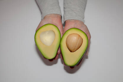Krupni plan ruku koje drže prepolovljeni avokado. Jedna polovina s košpom, druga bez nje, jasno otkrivaju glatku, kremastu, unutrašnjost. Tekstura avokada je svježa.