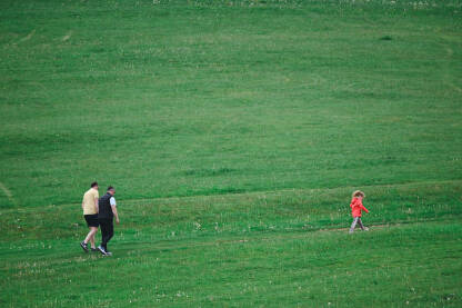 Dva čovjeka šetaju uskom stazom na livadi, dok je ispred njih djevojčica u crvenoj jakni koja gleda ispred sebe.