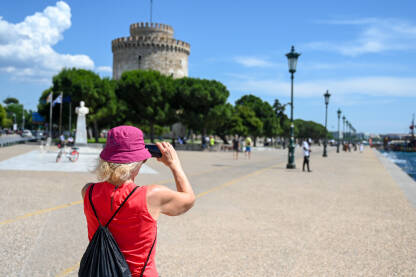 Žena fotografiše mobilnim telefonom u gradu. Žena fotografiše mobilnim telefonom u gradu. Turisti slikaju ulice i građevine. Šetalište u Solunu, Grčka.