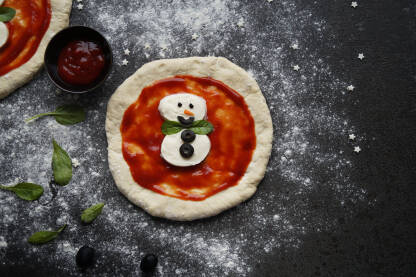 Snješko Bijelić Pizza Mala dječija pica u obliku Snješka Bijelića na na stolu sa brašnom, kečapom i crnim maslinama.