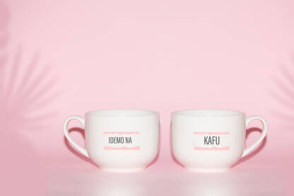 Bijele šolje na ružičastoj pozadini sa natpisom „Idemo na kafu“ .Moderan natpis za poslovni kućni ambijent.