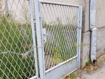 Lanac na kapiji Zatvorena kapija, učvršćena lancem i katancem, na ulazu u travom i korovom zaraslo dvorište; stara i napuštena kuća;