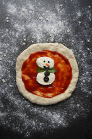 Snješko Bijelić Pizza Mala dječija pica u obliku Snješka Bijelića na na stolu sa brašnom, kečapom i crnim maslinama.
