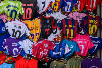 Štand sa velikim brojem sportskih dresova različitih boja i brojeva. Dresovi su okačeni na vješalice i gusto poredani jedan do drugog.