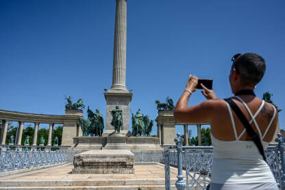 Budimpešta, Mađarska: Žena fotografira znamenitosti u gradu mobitelom. Turisti na Trgu heroja. Skulpture i spomenici. Grupa ljudi na trgu.
