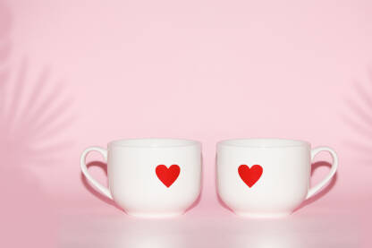 Dvije bijele keramičke šolje sa crvenim srcima stoje jedna pored druge na ružičastoj pozadini.Romantična kompozicija, za teme ljubavi.