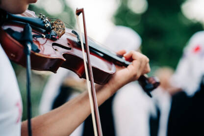 Muzičar svira violinu Folklor. Muzičar svira violinu. Violina ima smeđe tjelo i crni vrat, u pozadini folkloraši igraju kolo.