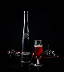 Vino od grožđa Elegantna flaša crnog vina, postavljene na stolu pored čaše sa crnim vinom i crnim grožđem u pozadini. Rubinovo crno/crveno vino Don Rossi.