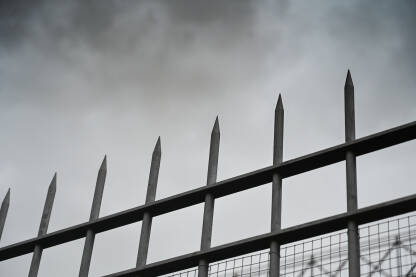Šiljci ograde uzdižu se prema olujnim oblacima. Vrh metalne ograde naspram tamnog dramatičnog neba. Oštra metalna ograda pod teškim tamnim oblacima.