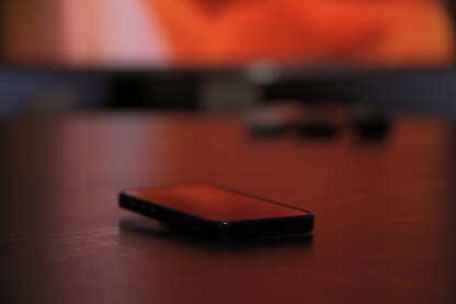 Mobilni telefon na stolu sa uključenim televizorom u pozadini.