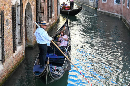 Gondola sa turistima u Veneciji Venecija, Italija: Gondola na rijeci. Istorijske građevine uz riječni kanal. Popularna turistička destinacija. Turisti istražuju grad.