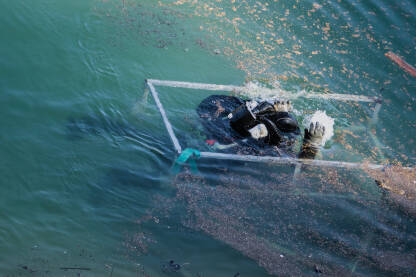 Ronilac izranja iz vode i ulazi u korpu za transport uz strmu obalu jezera. Ronioci, pod vodom, čiste naplavine materijala u jablaničkom jezeru.