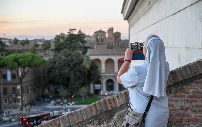 Časna sestra fotografiše grad Rim i Vatikan svojim mobilnim telefonom. Panoramski pogled na Rim i Vatikan tokom zalaska sunca. Kupole crkava. Siluete zgrada.