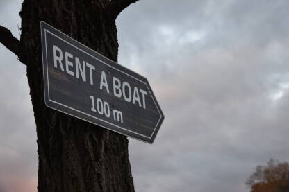 Rent a boat znak Znak za iznajmljivanje čamca na drvetu sa oblačnim nebom u pozadini.